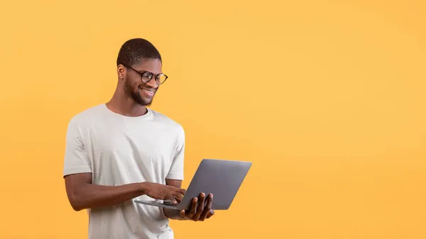 Ηλεκτρονική εκπαίδευση. Χαρούμενος Αφροαμερικάνος φοιτητής που κοιτάζει φορητό υπολογιστή και χαμογελαστό, κίτρινο φόντο, ελεύθερος χώρος — Φωτογραφία Αρχείου