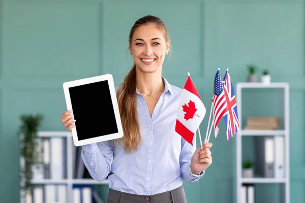 Šťastná učitelka ukazující podložku s prázdnou obrazovkou a držící vlajky různých zemí, pózující v úřadu — Stock fotografie