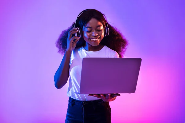 Genç siyahi kadın video görüşmesi yapıyor, dizüstü bilgisayar kullanıyor, neon ışıkta kablosuz kulaklık takıyor. — Stok fotoğraf