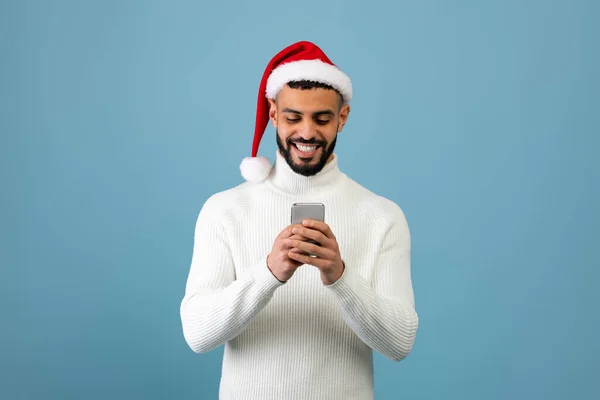 Vaya aplicación. Chico árabe feliz usando el teléfono celular usando sombrero de santa y sonriendo, de pie sobre fondo de estudio azul — Foto de Stock