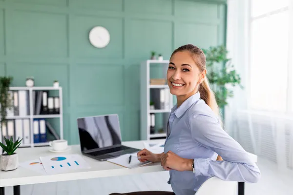 Succesvolle vrouwelijke ondernemer zittend aan bureau en laptop met blanco scherm in de buurt, kantoorinterieur, kopieerruimte — Stockfoto