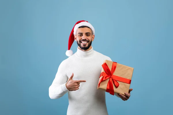 Entrega de regalos. Hombre árabe feliz en Santa sombrero sosteniendo caja regalo envuelto y señalando el dedo en él, fondo azul — Foto de Stock