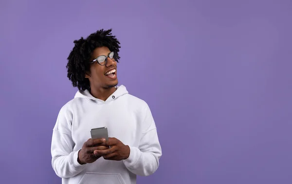 Feliz adolescente negro usando el teléfono inteligente, publicidad nueva aplicación, mirando a un lado en el espacio vacío sobre fondo violeta —  Fotos de Stock