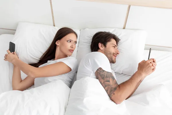 Perturbada esposa europeia ciumento e olha para o telefone do marido sorridente na cama branca no interior do quarto — Fotografia de Stock