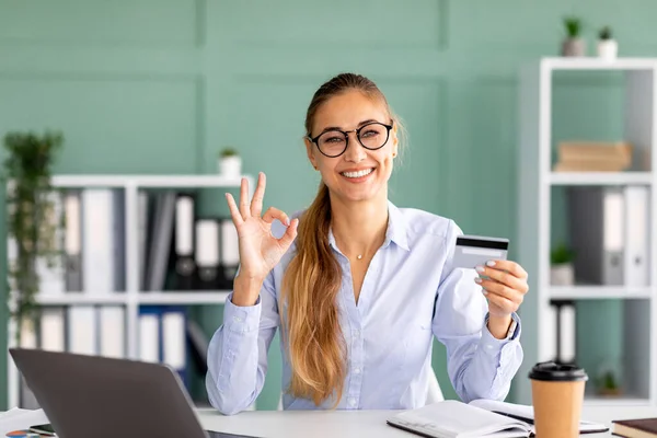 Boekhouder bankier dame met credit card en tonen ok teken tijdens het zitten op het werk, glimlachen op de camera in het kantoor — Stockfoto