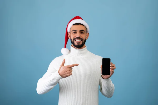 Vente de Noël en ligne. heureux arabe guy dans santa chapeau montrant smartphone avec écran vide et pointant sur elle, maquette — Photo