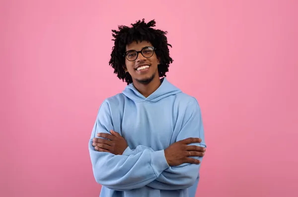 Portrét šťastný mladý černošky teenager v brýlích stojící se zkříženýma rukama, dívající se na kameru na růžovém pozadí — Stock fotografie