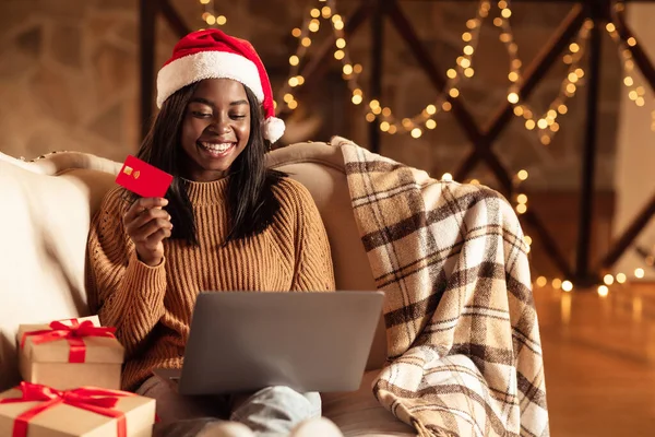 Online julshopping. Svart kvinna i Santa hatt sitter nära presentförpackningar, hålla kreditkort och använda laptop hemma — Stockfoto