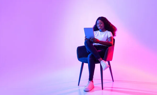 Joyeux dame noire utilisant une tablette, étudier ou travailler à distance, assis dans un fauteuil au néon, espace de copie — Photo
