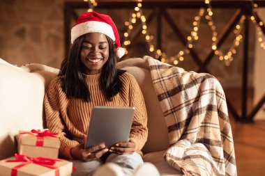 İnternetteki Noel kutlamaları. Noel Baba şapkalı siyahi kadın video çekiyor, tablet kullanıyor, Noel hediyesi alıyor.