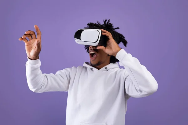 Niesamowite Afroamerykanin nastolatek facet w VR zestaw słuchawkowy za pomocą wirtualnej rzeczywistości dotykając wyimaginowany ekran na fioletowym tle — Zdjęcie stockowe