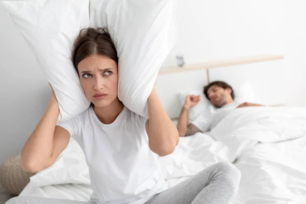 Уставшая недовольная кавказская жена закрывает уши подушкой, сидя на кровати со спящим мужем в спальне — стоковое фото