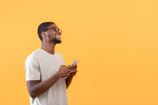 Oferta mobilna. Szczęśliwy Afroamerykanin trzyma smartfona i patrząc na wolne miejsce, żółte tło — Zdjęcie stockowe