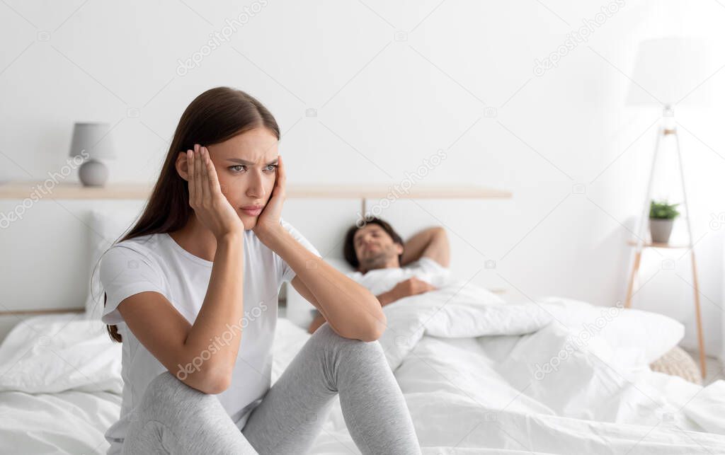 Sad offended european lady sitting on bed with sleeping husband in bedroom interior, free space