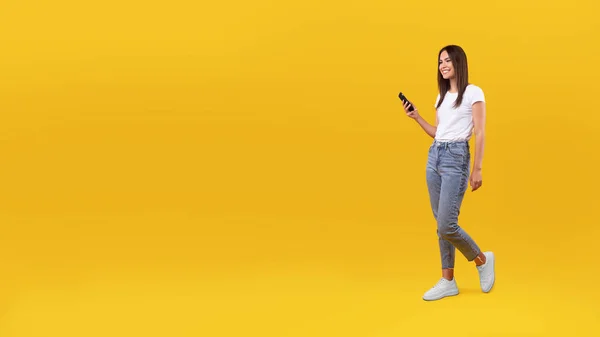 Sarı duvarda, panoramada cep telefonuyla yürüyen genç kadın. — Stok fotoğraf
