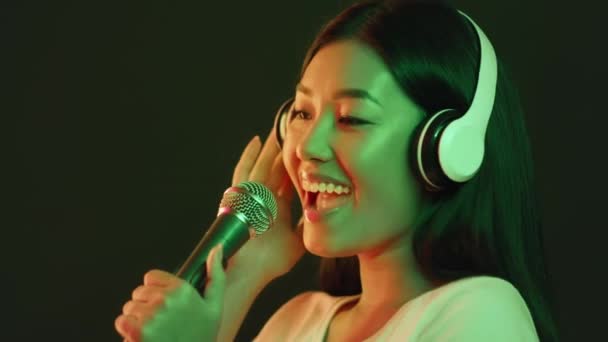 Молода азіатка-музикантка в професійних навушниках співає пісню на мікрофон на студії звукозапису в зелених неонових вогнях — стокове відео