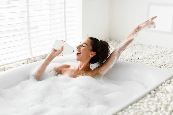 Mujer bastante joven tomando baño con espuma, usando botella de champú como micrófono, cantando canción favorita en casa —  Fotos de Stock