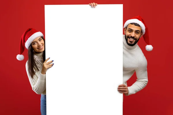 Reklama świąteczna. Szczęśliwa para arabska w kapeluszach Mikołaja trzymających białą tablicę, demonstrująca miejsce na tekst lub projekt — Zdjęcie stockowe