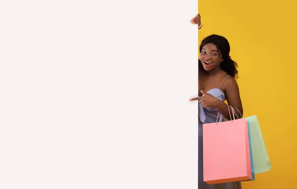 Jovem senhora preta feliz com sacos de compras apontando para banner de papel branco em branco com mockup para anúncio no fundo laranja — Fotografia de Stock