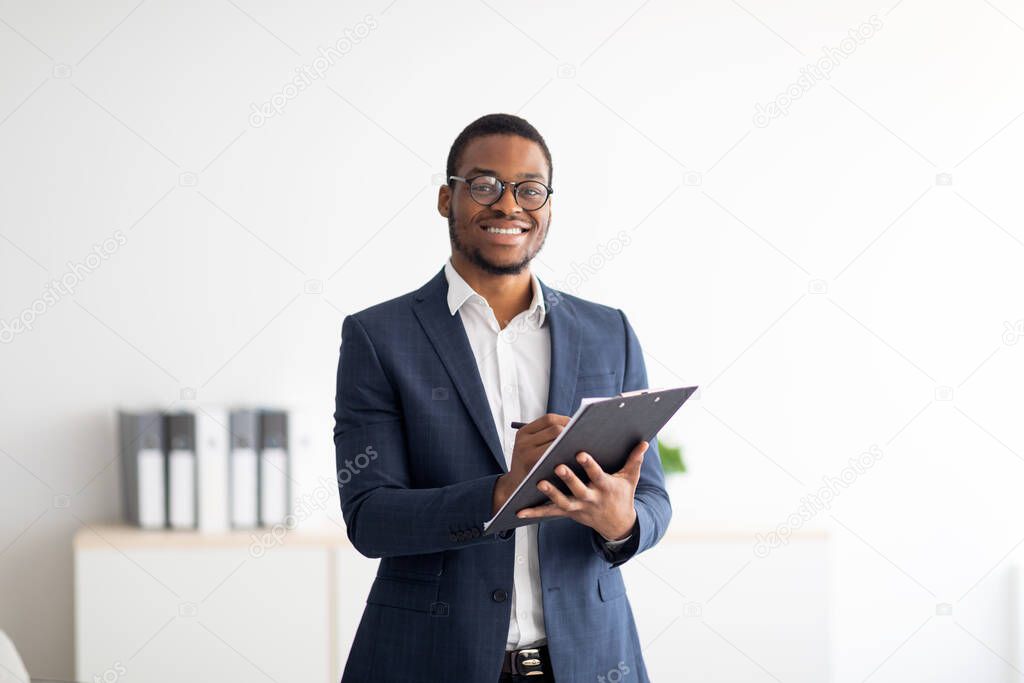 Happy young black male psychologist writing in clipboard, looking at camera and smiling in modern office