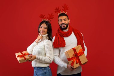 Geyik boynuzları içinde neşeli genç eşler ellerinde Xmas hediye kutuları ve kameraya gülümsüyor, kırmızı arka planda duruyorlar.