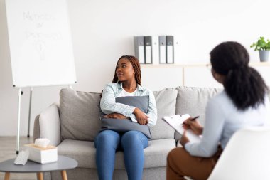 Sinir krizi geçiren siyah bir kadın psikologla seans yapıyor, koltukta oturuyor, klinikte yastığa sarılıyor.