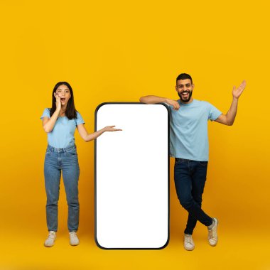 Şaşırmış Arap çift, sarı stüdyo arka planında boş ekranları olan akıllı telefonun yanında duruyor.