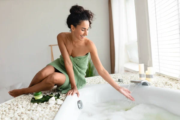 若い女性タオルでお風呂の近くに座って、水に触れ、温度をチェックし、国内のスパの手順の準備ができて — ストック写真