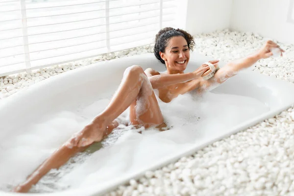 Jeune femme prenant un bain avec de la mousse, frottant sa peau soyeuse avec un pinceau à la maison. Hygiène, spa, concept relax — Photo