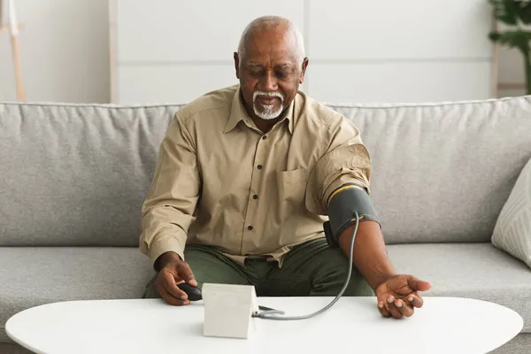 Masculino preto sênior medindo a pressão arterial com dispositivo de esfigmomanômetro em casa — Fotografia de Stock