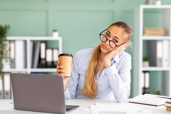 Retrato de jovem trabalhador de escritório cansado sentado no local de trabalho com laptop enquanto segurando xícara de café e olhando para a câmera — Fotografia de Stock