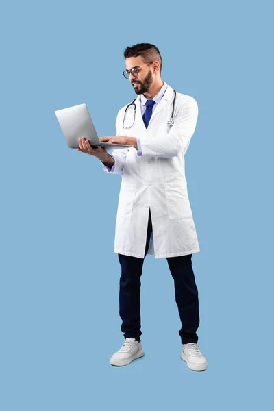 Arabo maschio medico utilizzando computer portatile in piedi su sfondo blu, verticale — Foto Stock