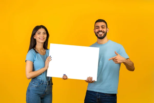Retrato de feliz pareja árabe sosteniendo cartel blanco, hombre apuntando al espacio libre para la publicidad, fondo amarillo —  Fotos de Stock