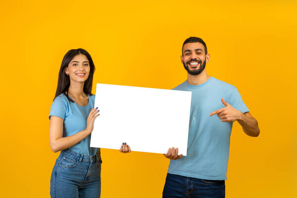 Portrait of happy arab couple holding white placard, man pointing at free space for advertising, yellow background