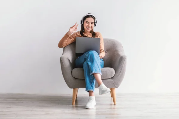 Mujer alegre que tiene llamada web o videoconferencia en el ordenador portátil, hablando en la cámara, usando auriculares, sentado en el sillón —  Fotos de Stock