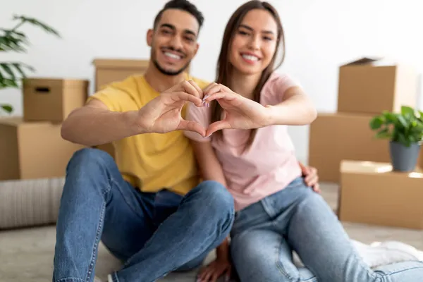 Cariñosa pareja multirracial haciendo un gesto de corazón con las manos, mudándose a una nueva casa, sentados en el suelo entre cajas —  Fotos de Stock