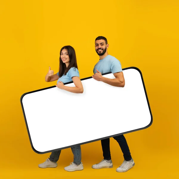 Heyecanlı Arap çift dizayn için büyük bir akıllı telefon tutuyor, sarı arka planda başparmağını gösteren kadın. — Stok fotoğraf