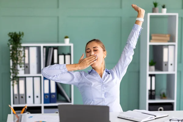 Trabalhador improdutivo. Jovem senhora bocejando e esticando as mãos enquanto sentado no local de trabalho no escritório moderno — Fotografia de Stock