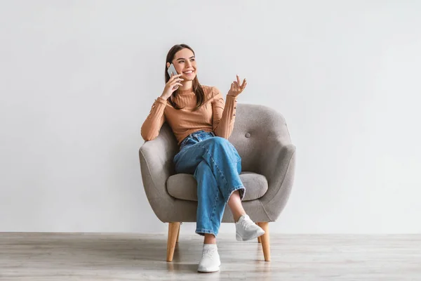 Mujer joven positiva hablando en el teléfono inteligente, haciendo llamada, sentado en sillón contra la pared del estudio blanco — Foto de Stock