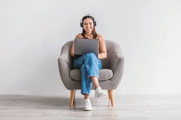 Χαρούμενη Καυκάσια κυρία που κάθεται στην πολυθρόνα με φορητό υπολογιστή και ακουστικά, επικοινωνώντας εξ αποστάσεως ή έχοντας επαγγελματική συνάντηση — Φωτογραφία Αρχείου
