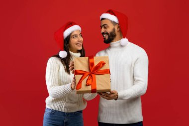 Sevgi dolu Arap çift Noel hediyesi kutusunu tutuyorlar ve birbirlerine gülümsüyorlar, Noel Baba şapkaları takıyorlar, kırmızı arka planda poz veriyorlar.