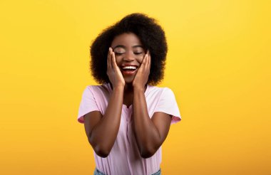 Mutluluk ve neşe. Güzel Afro-Amerikalı kadın yanaklarından elele tutuşuyor ve gözleri kapalı gülümsüyor, sarı arka plan