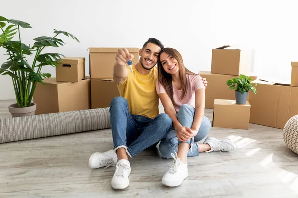 Portret pełnej długości kochającej się międzyrasowej pary siedzącej na podłodze wśród kartonowych pudełek, pokazującej klucz do domu w pomieszczeniu — Zdjęcie stockowe