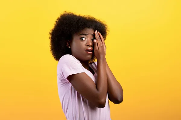 Jovem senhora negra estressada cobrindo rosto com as mãos e espreitando com um olho, de pé sobre fundo amarelo — Fotografia de Stock