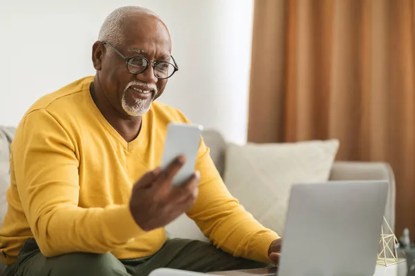 Homem preto sênior usando Smartphone e laptop trabalhando on-line no interior — Fotografia de Stock