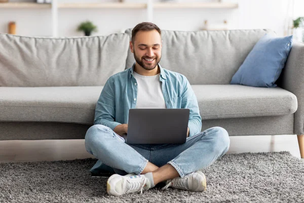 Full length of happy Καυκάσιος άνδρας κάθεται σταυροπόδι στο πάτωμα, χρησιμοποιώντας φορητό υπολογιστή, που εργάζονται σε απευθείας σύνδεση από το γραφείο στο σπίτι — Φωτογραφία Αρχείου