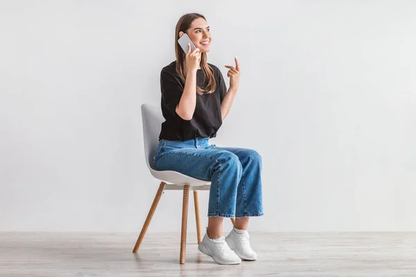 Sonriente joven mujer teniendo conversación en el teléfono celular, sentado en una silla acogedora contra la pared del estudio blanco, longitud completa —  Fotos de Stock