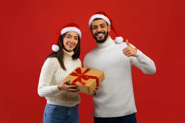 Happy young arab couple in Santa hats holding gift box and man showing thumb up, smiling to camera, red background — Stock Photo, Image