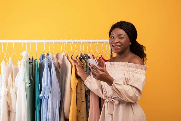 Siyah kadın internet alışverişi için cep telefonu kullanıyor, elbise rafının yanında duruyor, turuncu arka planda yeni kıyafetler seçiyor. — Stok fotoğraf
