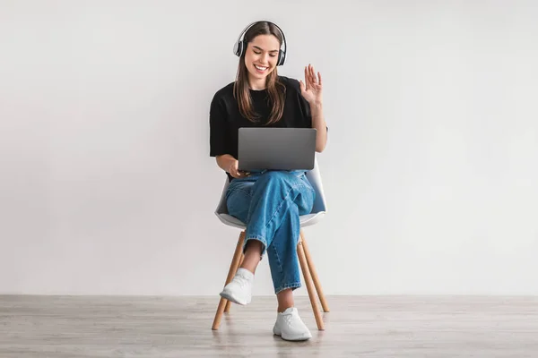 Χαρούμενη νεαρή γυναίκα με ακουστικά που χαιρετά την κάμερα του φορητού υπολογιστή, συνομιλεί στο διαδίκτυο, έχει τηλεδιάσκεψη, κάθεται στην καρέκλα — Φωτογραφία Αρχείου